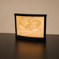 A black rectangular photo lamp or night light sits on a dark wooden table against a warm beige wall. The illuminated front displays a softly glowing sepia-toned image of a woman kissing a sleeping baby.