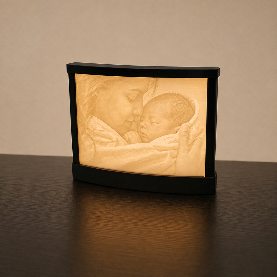 A black rectangular photo lamp or night light sits on a dark wooden table against a warm beige wall. The illuminated front displays a softly glowing sepia-toned image of a woman kissing a sleeping baby.