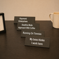 Four rectangular black desk nameplates with white text are arranged in a staggered row on a dark wood table. A cream mug is partially visible on the right, and a black frame or photo frame edge is partially visible on the left.