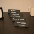 Four rectangular black desk nameplates with white text are arranged in a staggered row on a dark wood table. A cream mug is partially visible on the right, and a black frame or photo frame edge is partially visible on the left.