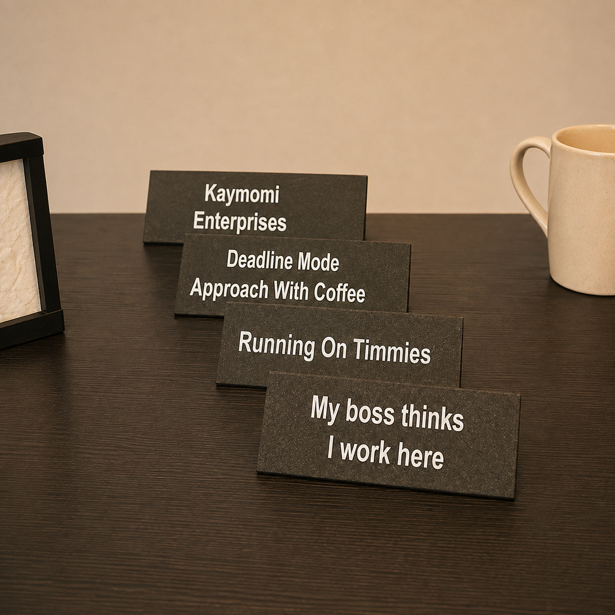 Four rectangular black desk nameplates with white text are arranged in a staggered row on a dark wood table. A cream mug is partially visible on the right, and a black frame or photo frame edge is partially visible on the left.