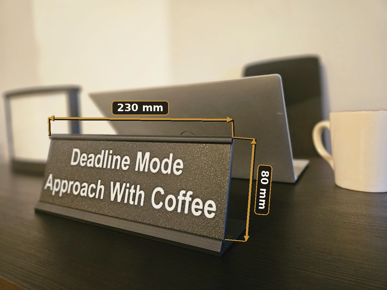 A dark gray desk sign with white text reading 'Deadline Mode Approach With Coffee' on a dark tabletop, with overlaid dimension labels showing 230 mm width and 80 mm height. A laptop and white mug are blurred in the background.