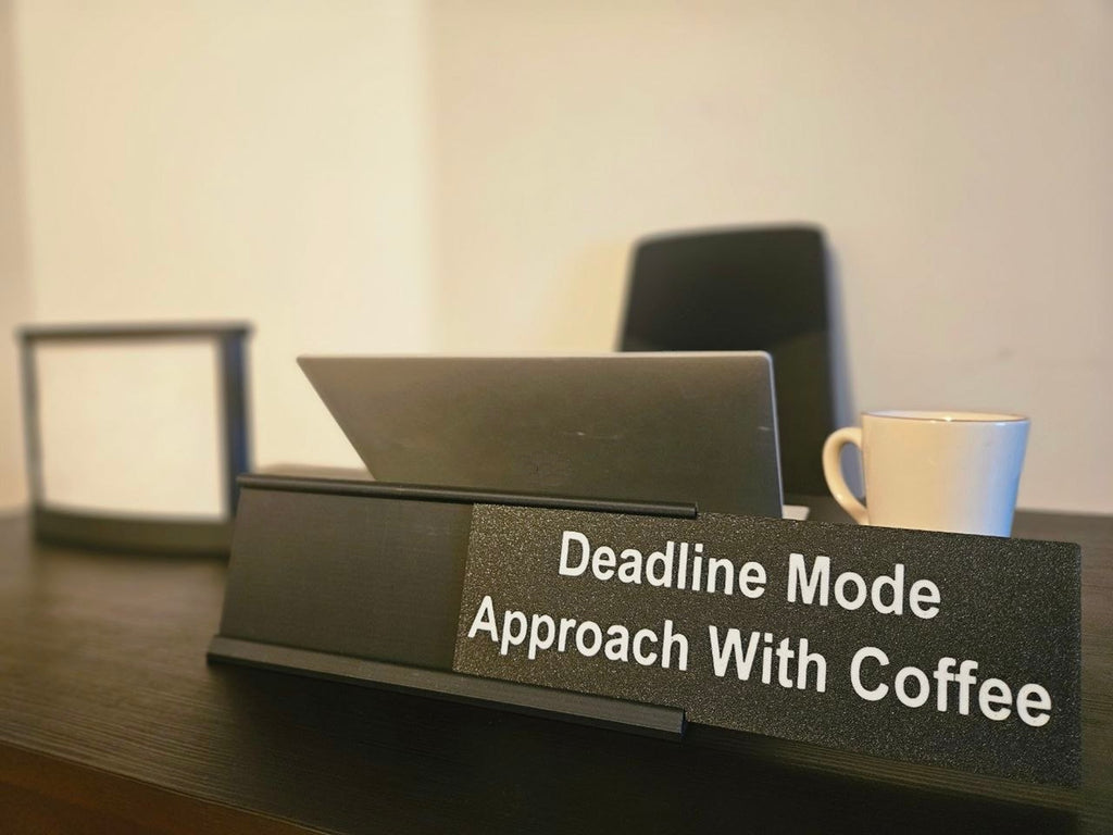 A black desk nameplate with the text 'Deadline Mode Approach With Coffee' in white lettering, shown on a dark wooden desk with a laptop and a white mug in a softly lit office setting.