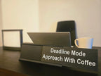 A black desk nameplate with the text 'Deadline Mode Approach With Coffee' in white lettering, shown on a dark wooden desk with a laptop and a white mug in a softly lit office setting.