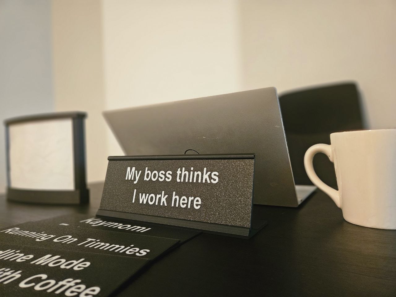 A tabletop scene with a small black sign reading 'My boss thinks I work here' in white text, alongside other humorous desk signs, a laptop, and a white mug on a dark table.