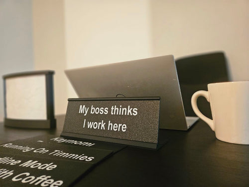 A tabletop scene with a small black sign reading 'My boss thinks I work here' in white text, alongside other humorous desk signs, a laptop, and a white mug on a dark table.