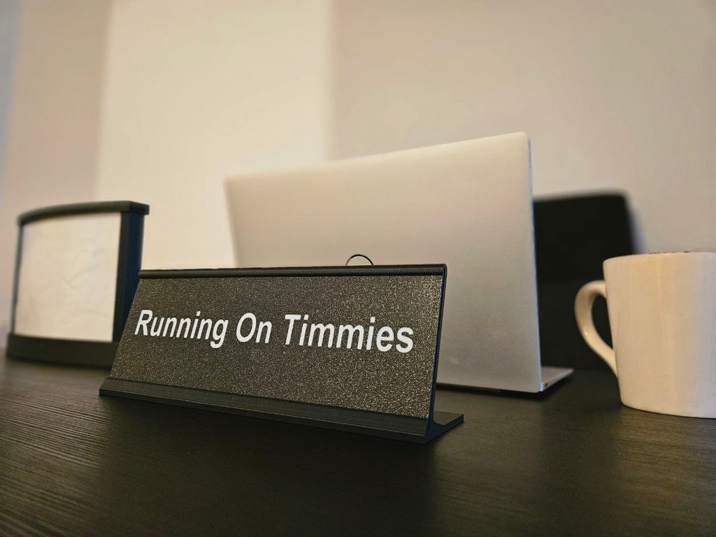 A black textured desk nameplate with white text reading 'Running On Timmies' standing on a dark wooden table, with a laptop, mug, and lamp blurred in the background.