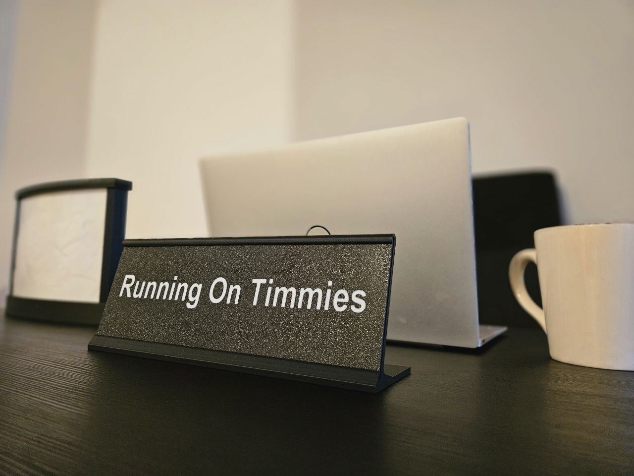 A black textured desk nameplate with white text reading 'Running On Timmies' standing on a dark wooden table, with a laptop, mug, and lamp blurred in the background.